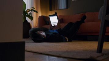 giovane asiatico sul tappeto, parte inferiore delle gambe sul sedile del divano, cuffia sulle orecchie, tenendo il tablet video