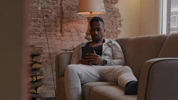 Mature man sits on couch watching tablet, leans back on armrest, stretched leg on couch, other hand touches head, upper body moves slightly video