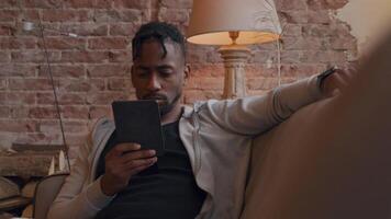 Mature man sits on sofa, watching and holding tablet with one hand in front of him. Other hand on backrest, having a look on his smartwatch, talks shortly, looks at smartwatch again video