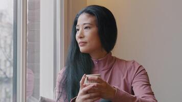 Mature woman sits on window sill, keeps looking through window and holds cup in both hands, takes sip with cup in one hand, holds cup in both hands again. video