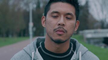 Close-up of young Asian man, standing outside, looking into camera, turns head to the right camera, looking back into camera, putting on face mask, adjusting it. Slow motion video