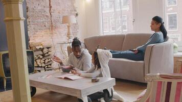 homem e menina sentada no chão à mesa. livro e lápis de cor na frente deles. falam, gesticula o homem, olha a menina e a menina olha o homem. ambos começando a desenhar no livro de trabalho. com lápis. mulher senta com as pernas esticadas no sofá, assiste televisão, be ... video