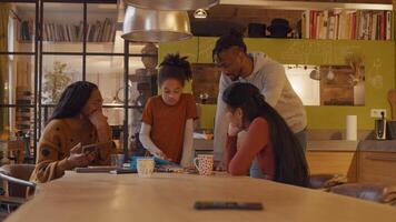 Girl stands in front of table watching and scrolling on children tablet. Two women, sitting around table and man standing, all watching tablet. Woman on the left - camera - talks with girl, points at tablet, girl talks, scrolls, taps and gestures. video