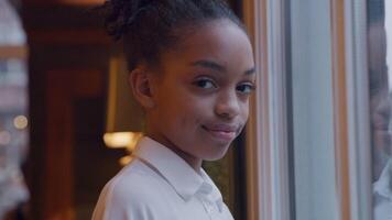 Black girl stands in front of window, head and eyes turned to camera, looking into camera lens, smiling slightly video