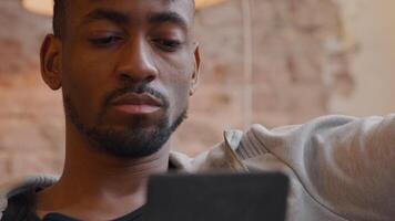Mature black man holds tablet in front of him, hand touching head twice. Blurred tablet video