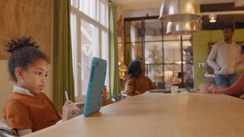 Girl sits at table, holds tablet with one hand in front of her, stylus pen in other hand. In background mature man standing and two mature women sitting at table, talking. video