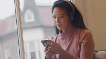 Mature woman sits on window sill, headphone on ears, slightly moving her head, holding cell phone in front of her, watching it
