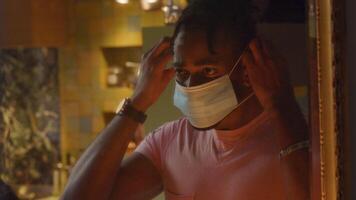 Mature black man in front of mirror, looks down, looks up, puts on face mask, whilst looking into mirror. Adjusts mask.Face is seen in mirror. Blurred doorway on the left camera video