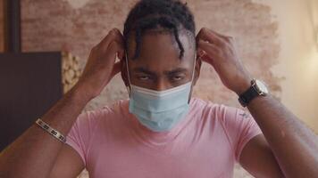 Mature black man turns head down, puts on face mask, looking into camera lens, adjusting mask and leaves video