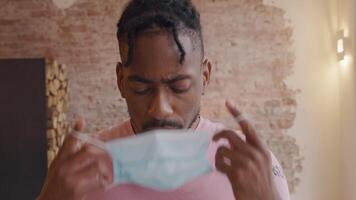 Mature black man looks serious, contemplative, searching eyes. He looks down, puts on face mask and looks around video