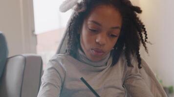 Black girl sits at table with work book. Holds pencil whilst writing in book. Talks. video