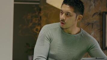 Young Asian man, whilst busy tapping on screen of device, he talks, turns his head,eyes to the left and back, frowning his eyebrows and forehead repeatedly video