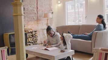 hombre y niña sentados en el suelo a la mesa. libro y crayones delante de ellos. el hombre habla seriamente con la niña, hace gestos, mira a la niña. la niña lo mira, dibuja en el libro, el hombre sonríe, le pone la mano en los hombros, asiente y él también comienza a dibujar. mujer sentada con ... video