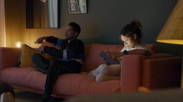 Asian young man and black young woman sitting on couch, man watches television, uses remote control and woman watches tablet . video