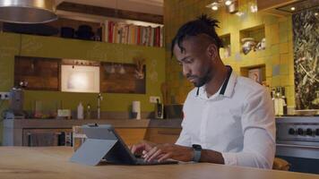 Mature man sits at kitchen table, typing on his laptop, watches on smartphone, tapping on it video