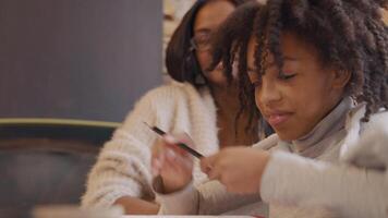 Woman and girl sit at table. Girl chooses colored pencil, reads in work book in front of her. Woman points at book, index finger following lines, whilst taking to girl. video