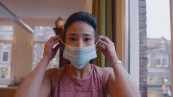 Mature Asian woman looks through window,profile, turns to the camera, looks in camera lens, puts on face mask, adjusts it and leaves video