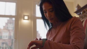 Mature Asian woman sits in armchair, looks through window. Looks on smartwatch, whilst moving arm up.zoom Swipes on screen of smartwatch video