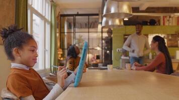 Girl sits at table, holds tablet with one hand in front of her, stylus pen in other hand. In background mature man standing and two mature women sitting at table, talking. video