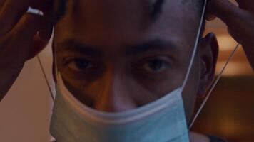 Zoom, close up of black mature man, looking into camera lens, putting on face mask, adjusting it, turns head to window light, looks back into camera video