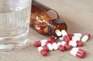 Cápsulas de pastillas rojas y blancas y vidrio en botella vertiendo sobre mesa de madera