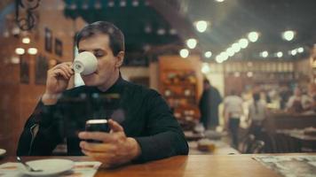 joven empresario tomando café y escribiendo sms en un café. Ver a través del cristal con débil reflejo de la ciudad. video