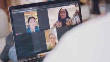 Close up of laptop screen with video call with three women in three frames shown. Laptop on lap of person who gestures.