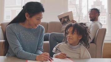 Woman and girl sitting on floor at table. Book and crayons in in front of them. They talk, woman points with pencil at work book. Girl  smiles, talks, pointing with pencil on work book too. Man on couch in background, reads paper, watches girl, laughs... video