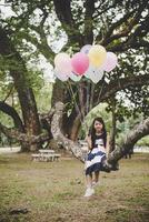 Linda niña asiática sentada en la rama de un árbol con globos de colores foto