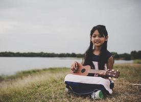 Linda niña asiática sentada en el césped y tocando el ukelele en el parque