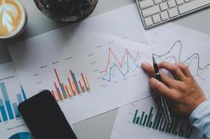 La mano de un hombre de negocios sosteniendo un bolígrafo sobre documentos comerciales, gráficos, informes e inversiones en una mesa gris, teléfono móvil, café y teclado de computadora. foto