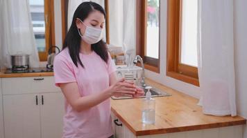 uma mulher usando uma máscara limpa as mãos com álcool gel na cozinha