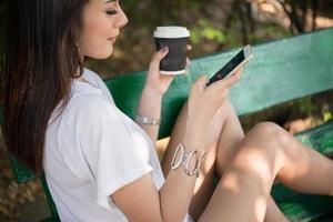 Retrato de una mujer casual feliz sentada en un banco con café y teléfono en el parque