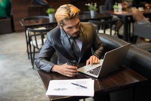 Young businessman holding cup of coffee while working on laptop computer in coffee shop