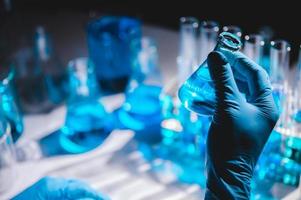 Hand in blue glove holding flask of blue liquid with flasks and vials of blue liquid in the background photo