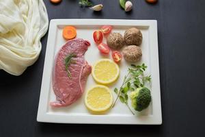 Filete crudo, perejil, champiñones, zanahorias y tomates en rodajas en un plato blanco sobre la mesa de madera negra foto