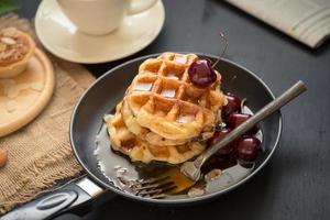 Waffles and cherries with honey in a pan, crispy almond tarts, and a cup of coffee on black table photo