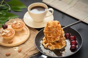 gofres y cerezas con miel, tartas de almendras crujientes, taza de café y un periódico doblado sobre una mesa negra foto