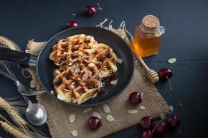 Miel en waffles en una sartén con cerezas sobre mesa de madera oscura. foto
