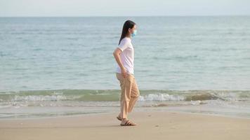 mujer asiática con mascarilla protectora caminando por la playa.