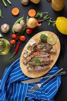 Grilled steak and garnishes on wooden cutting board with onions, chili peppers, tomatoes, green beans, and a lemon on dark wooden table photo