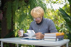 Hombre joven barba hipster tomando café mientras lee libros en el jardín de su casa con la naturaleza foto