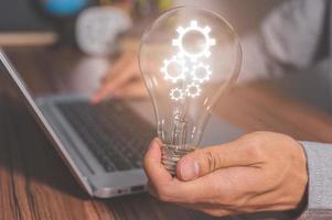 A man holding a light bulb gear symbol while using a computer photo