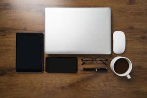 Top view of workspace with laptop, smartphone, tablet, coffee cup, glasses and pen on wooden table photo