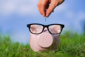 Pink piggy bank with glasses on grass and hand putting in a coin photo