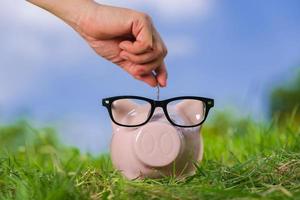 Pink piggy bank with glasses on grass and hand putting in a coin photo