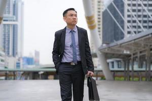 Asian businessman walking and holding briefcase with business office buildings in the city background photo
