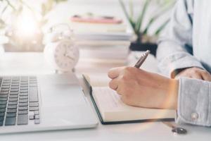 Woman writing in a planner notebook while using laptop at home photo