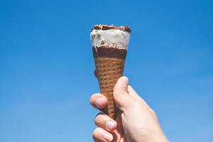 Hand holding a cone of ice cream with a blue sky background, summer concept
