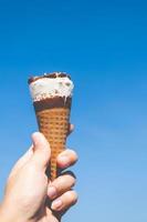 Hand holding a cone of ice cream with a blue sky background, summer concept